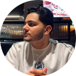 Foto de Guilherme Turra em um restaurante, com roupa branca e uma logo no meio, olhando para o lado esquerdo.